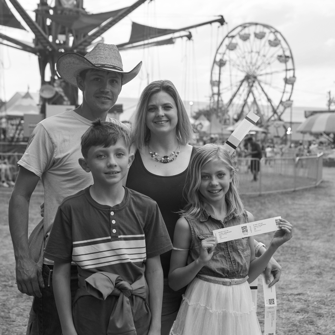 This black and white photo captures a happy family at the fair, posing with their tickets in hand. The father, wearing a cowboy hat and t-shirt, stands proudly next to the mother, who smiles warmly in a sleeveless top and necklace. Their two children, a boy and a girl, stand in front, excitedly holding their fair tickets. The girl wears a denim top and a white skirt, while the boy sports a casual striped shirt with a sweater tied around his waist. In the background, the bustling fairground features a Ferris wheel and various rides, creating a fun and lively atmosphere perfect for a family day at the fair.