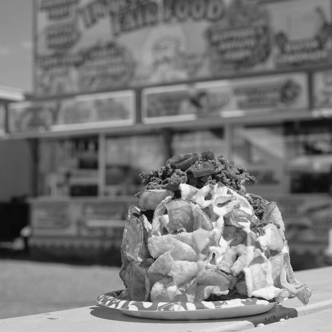 A black and white photograph showcases a towering plate of loaded fair food, featuring crispy, golden ribbon fries drizzled with creamy ranch, nacho cheese sauce and topped with seasoned carne asada, jalapeños, and other savory toppings. The dish is placed on a picnic table in the foreground, highlighting its mouth-watering texture and detail. In the slightly blurred background, a bustling fair food stand with large, colorful signage advertises various treats, with flags fluttering in the sky above. The image captures the indulgent and festive atmosphere of enjoying classic fair cuisine.