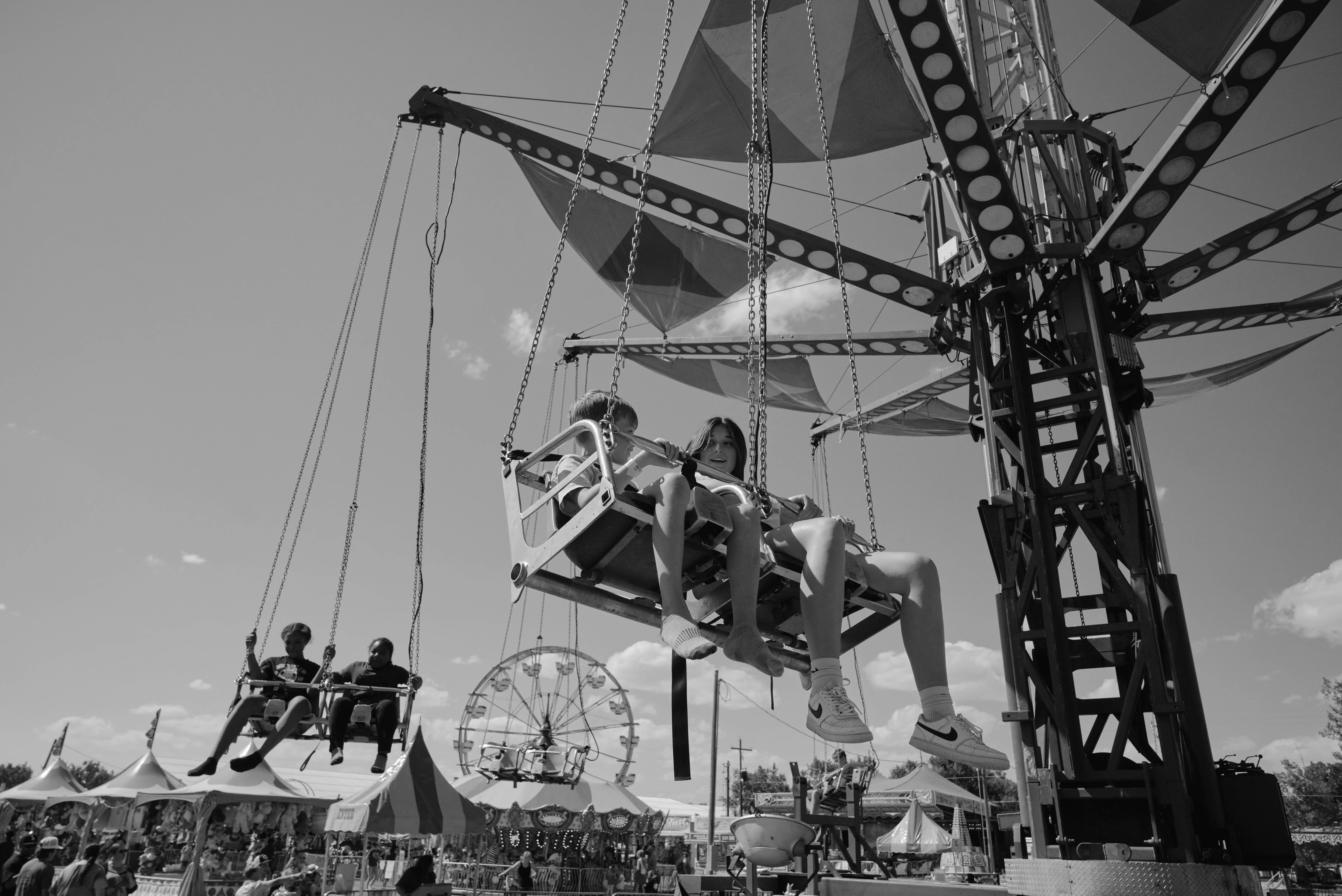 B&W Swings Ride A black and white photograph captures a thrilling moment at a fair as children enjoy a swing ride under a clear sky. Two kids sit in the foreground, suspended mid-air by sturdy chains, with expressions of excitement and anticipation. Their legs dangle freely, with one wearing sneakers and the other in socks, adding to the carefree atmosphere. In the background, another pair of riders can be seen, along with a Ferris wheel and various fair tents bustling with activity. The ride's structure features bold geometric patterns and sturdy beams, framing the joyful scene with a mix of nostalgia and excitement characteristic of classic fair experiences.