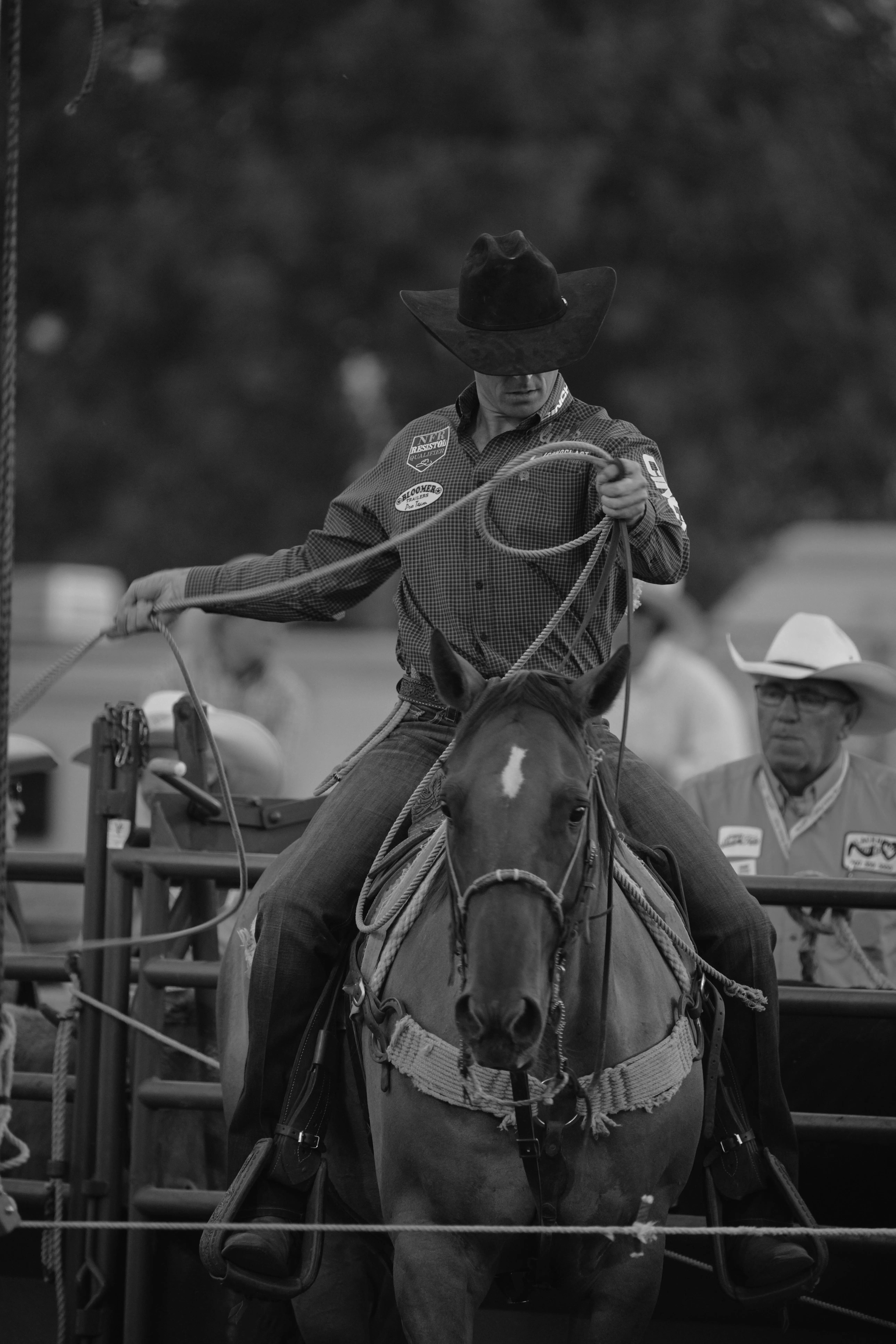 B&W Horseback Cowboy A black and white photograph captures a cowboy preparing for a rodeo event. He is seated on a well-groomed horse, holding a lasso in his right hand, ready to make his move. The cowboy is wearing a checkered button-up shirt adorned with sponsor patches, jeans, and a black cowboy hat that shades his eyes, giving him a focused and determined look. His horse, outfitted with a bridle and sturdy saddle, appears calm and attentive. In the background, another cowboy wearing a white hat and a Western-style shirt observes the scene, adding depth and context to the rodeo setting. The composition of the image emphasizes the intensity and tradition of rodeo culture.