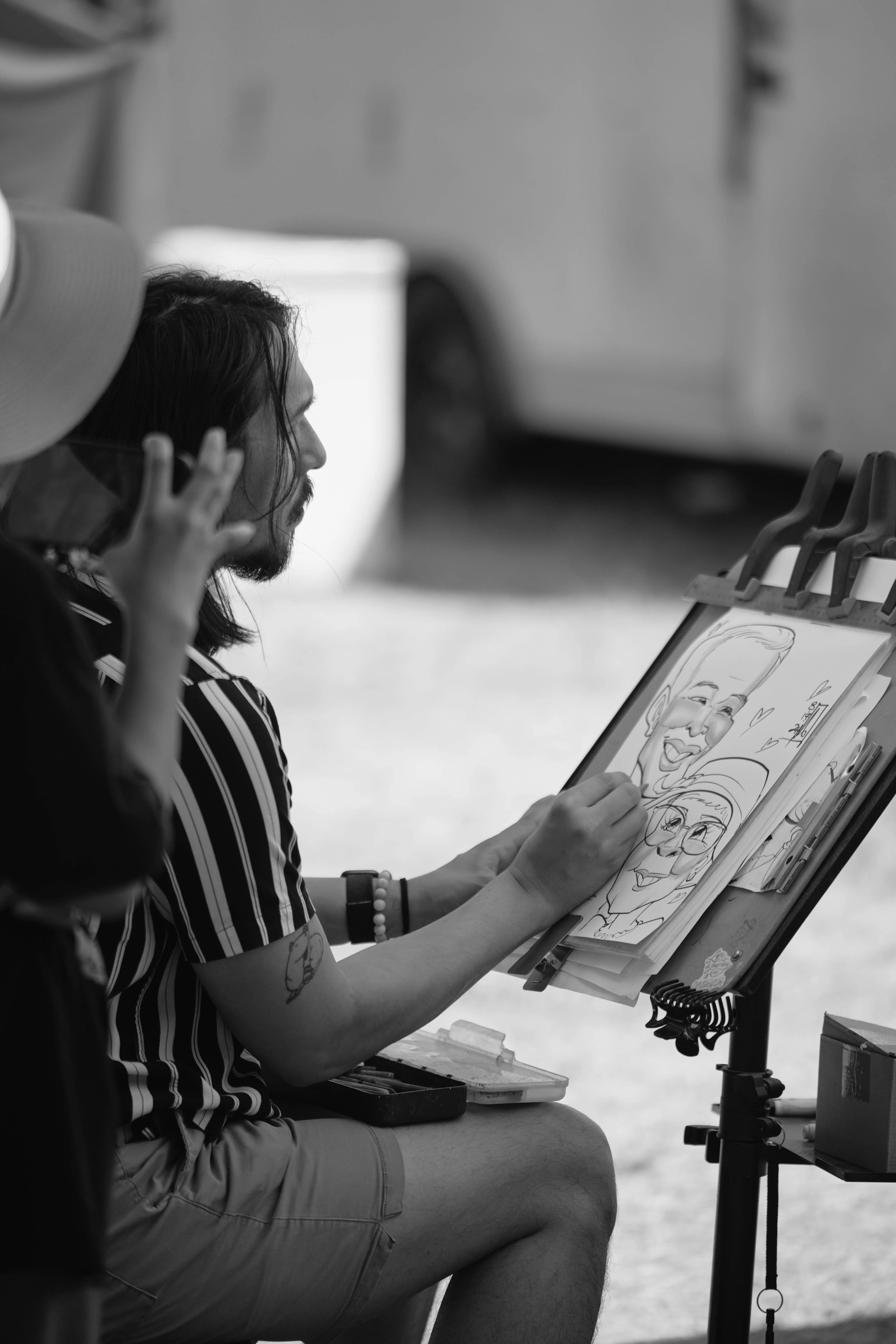 B&W Caricature This black and white photo captures a talented caricature artist at work during the fair. Seated with a focused expression, the artist skillfully brings a lighthearted portrait to life, exaggerating the features of a smiling couple on the drawing pad. The subject, wearing a sun hat, watches closely, capturing the fun and creativity of the moment. Various drawing tools and supplies are neatly arranged on the artist's easel, adding to the professional setup. In the background, fair tents and activity provide context to this engaging scene, showcasing the lively and artistic atmosphere of the fair.