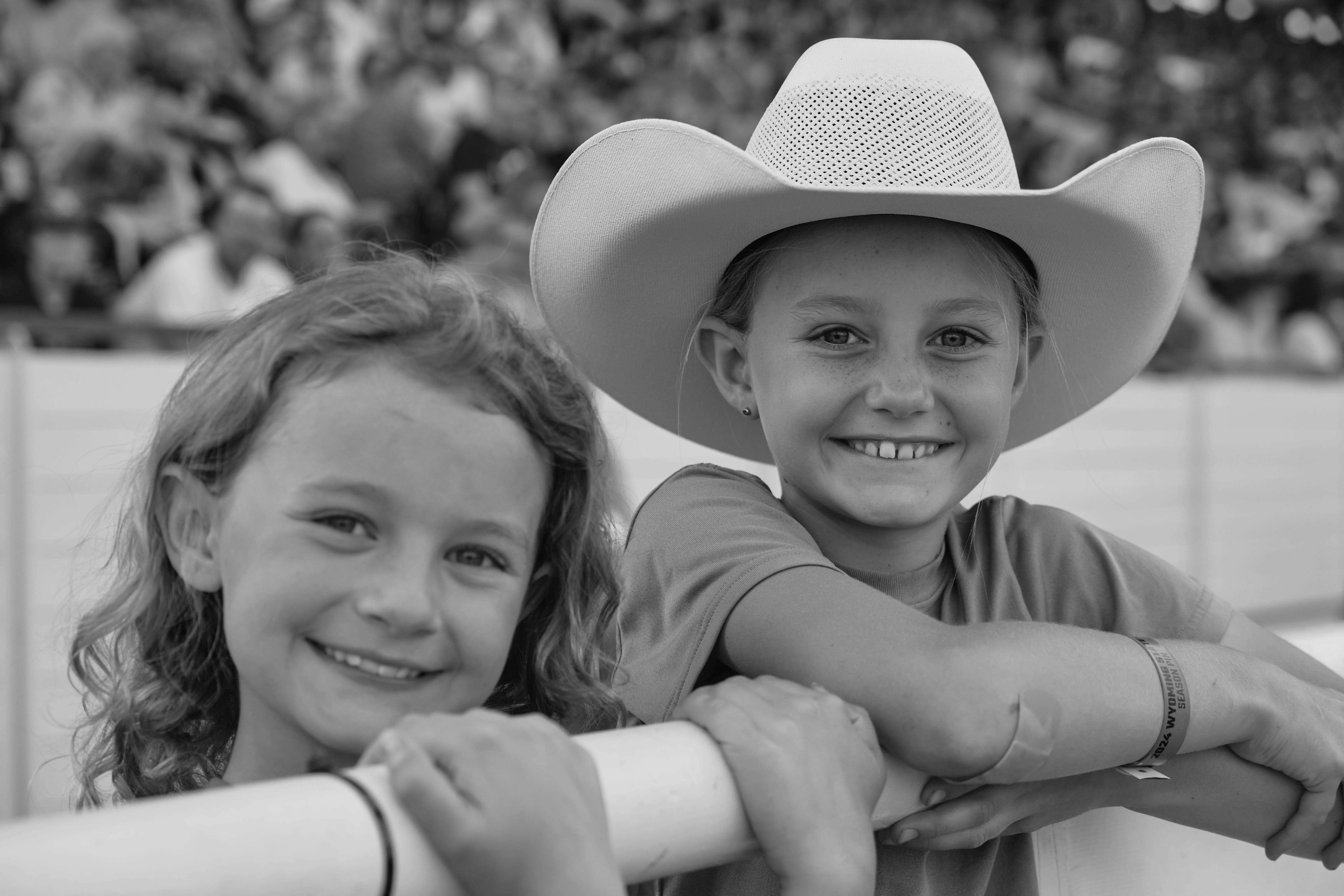 B&W Cowgirl Kids This black and white photo captures the joy and excitement of two young fairgoers leaning against a white fence, smiling brightly. One girl, wearing a classic cowboy hat and a wristband from the Wyoming State Fair, exudes Western charm and enthusiasm, while her friend, with curly hair and a beaming smile, radiates happiness. The blurred background shows a crowd of fair attendees, adding to the lively atmosphere of the event. This image beautifully showcases the spirit of the fair—bringing together families, friends, and communities to create unforgettable memories filled with laughter and Western traditions.