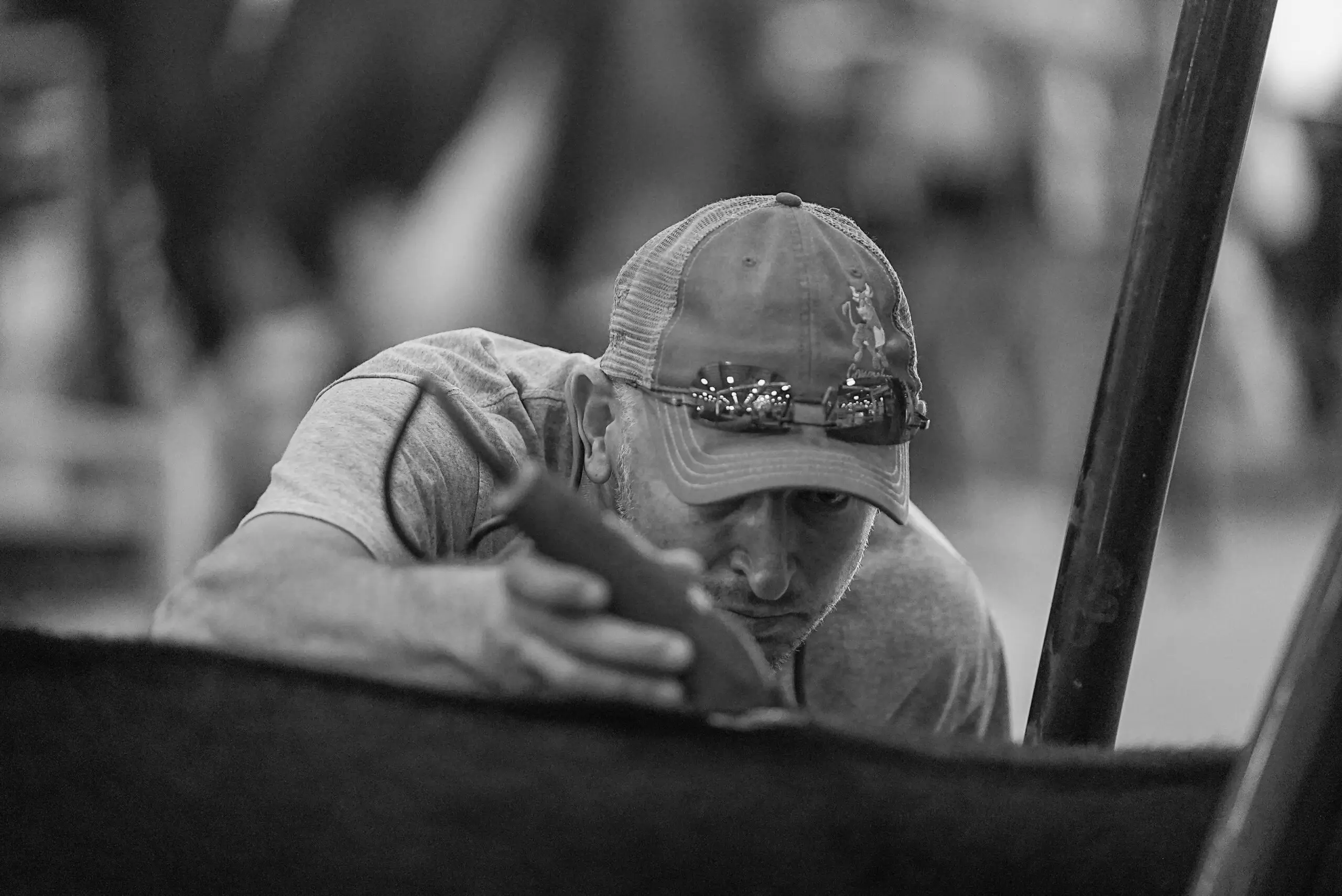 B&W Fitting This black and white photo captures a livestock fitter meticulously grooming a steer in preparation for the show ring. Focused and determined, he carefully trims the animal's coat with precision, ensuring it looks its best for the judges. Wearing a cap with sunglasses resting on the brim, his attention to detail reflects the dedication and craftsmanship that goes into preparing show cattle. The blurred background of the barn captures the bustling atmosphere of the fair, where exhibitors and fitters work tirelessly behind the scenes. This image highlights the artistry and commitment that contribute to the competitive spirit of livestock showing.