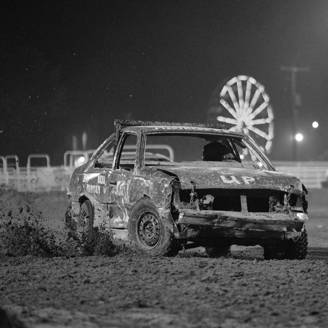 This black and white photo captures the excitement of a Figure 8 Race car at the fair. A battered car, covered in dents and mud, powers through the dirt track with its front end missing parts, showing the wear and tear of the intense competition. The driver, visible through the open window, is focused and determined. In the background, the bright lights of the fairground and a Ferris wheel add to the atmosphere, creating a perfect blend of adrenaline and nostalgia. Dirt flies from the car's tires, emphasizing the high-energy action of this crowd-favorite event.