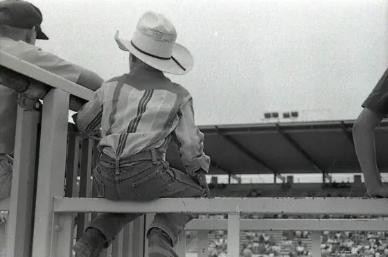 A black and white photograph captures a cowboy preparing for a rodeo event. He is seated on a well-groomed horse, holding a lasso in his right hand, ready to make his move. The cowboy is wearing a checkered button-up shirt adorned with sponsor patches, jeans, and a black cowboy hat that shades his eyes, giving him a focused and determined look. His horse, outfitted with a bridle and sturdy saddle, appears calm and attentive. In the background, another cowboy wearing a white hat and a Western-style shirt observes the scene, adding depth and context to the rodeo setting. The composition of the image emphasizes the intensity and tradition of rodeo culture.