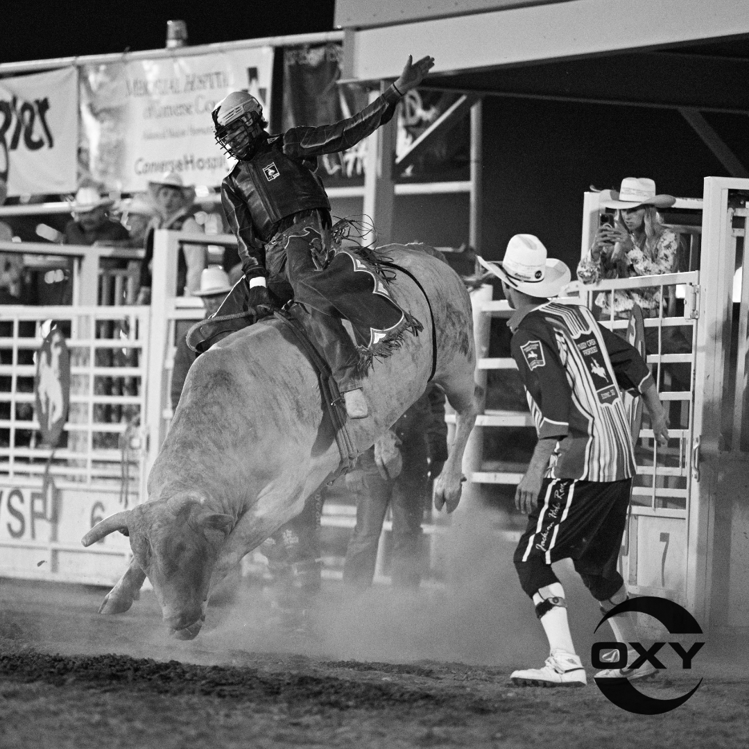 A black and white photo captures an exciting moment at a rodeo as a bull rider hangs on tight to a powerful bucking bull. With one arm raised for balance, the rider focuses on staying on while the bull kicks up dust, showing off its strength. A rodeo clown in a striped shirt and cowboy hat stands nearby, ready to jump in and help if needed. In the background, fans and event staff watch closely, with some snapping photos and others cheering. This image perfectly captures the thrill, energy, and tradition of rodeo life.