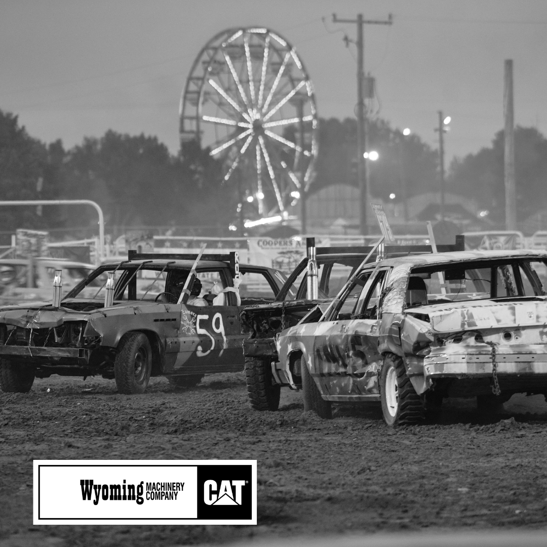 This black and white photo captures the excitement of a demolition derby at the fair, with a row of battered cars lined up in the dirt arena, their dented bodies showing signs of previous collisions. The number "59" is boldly painted on one of the vehicles, standing out against the rough, worn exterior. In the background, the glowing lights of a Ferris wheel illuminate the night sky, adding a festive touch to the gritty competition. Power lines and fairground structures frame the scene, emphasizing the blend of adrenaline-pumping action and classic fair nostalgia.