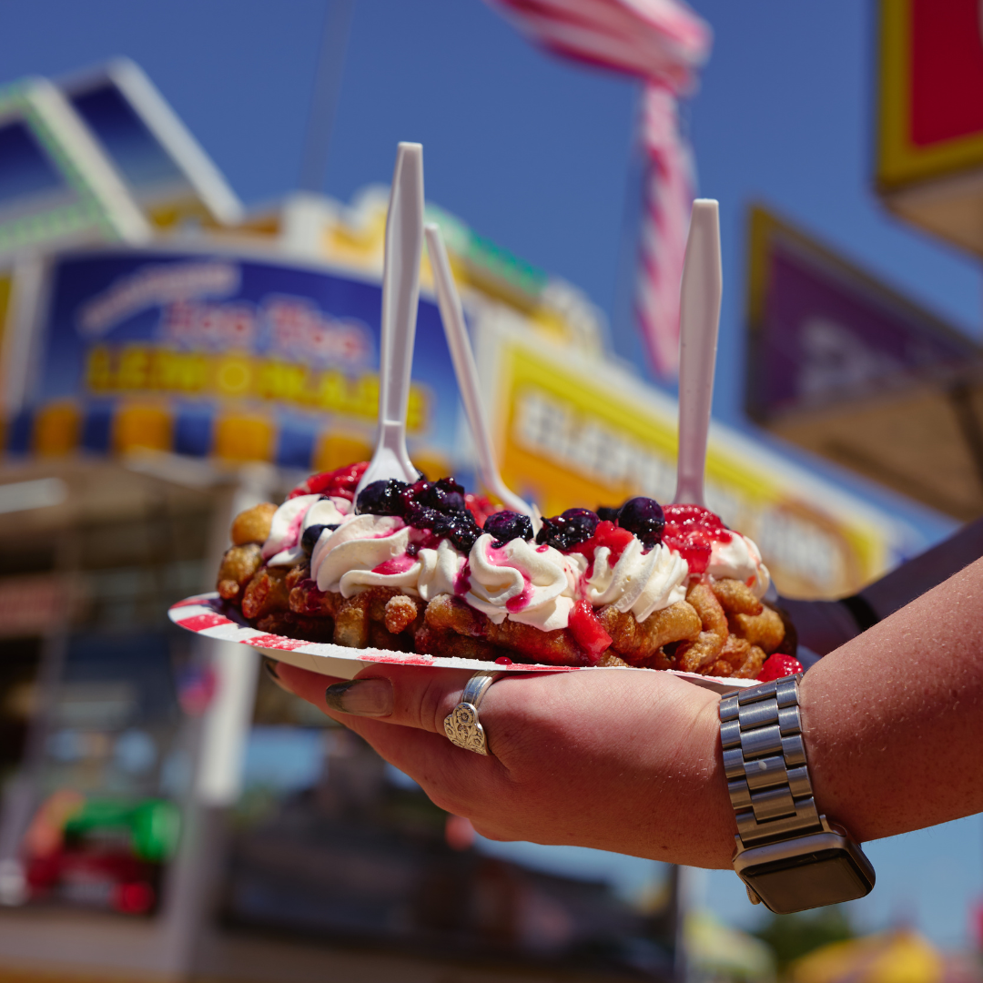 Funnel cake topped with whipped cream and berries held in front of a fair food stand at the Wyoming State Fair.
