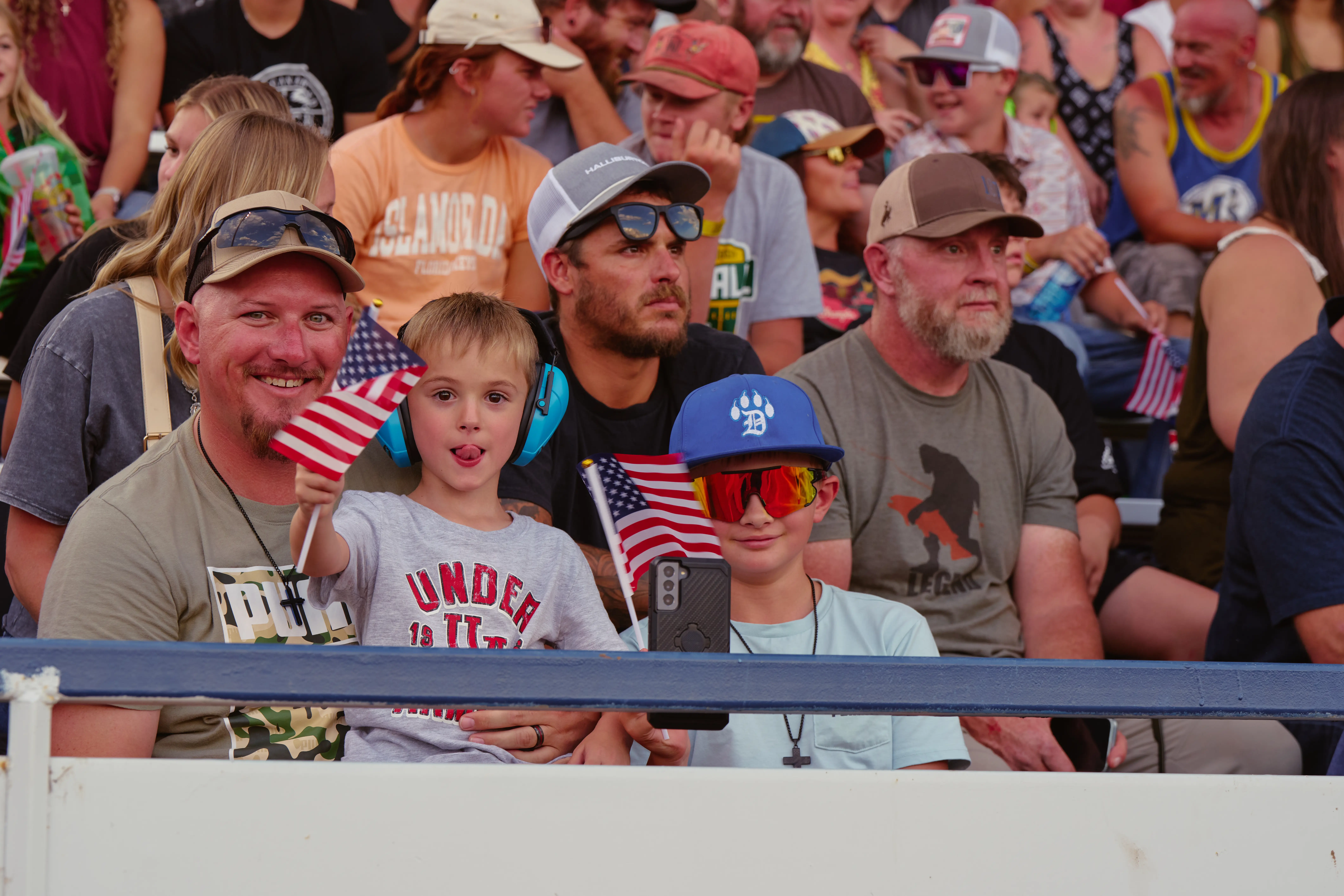 This black and white photo captures the joy and excitement of two young fairgoers leaning against a white fence, smiling brightly. One girl, wearing a classic cowboy hat and a wristband from the Wyoming State Fair, exudes Western charm and enthusiasm, while her friend, with curly hair and a beaming smile, radiates happiness. The blurred background shows a crowd of fair attendees, adding to the lively atmosphere of the event. This image beautifully showcases the spirit of the fair—bringing together families, friends, and communities to create unforgettable memories filled with laughter and Western traditions.