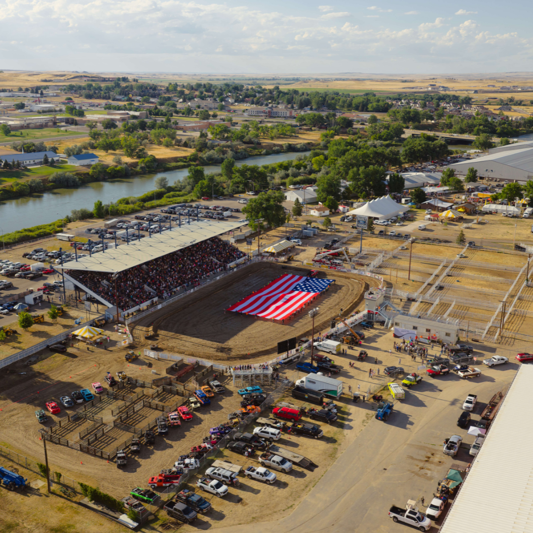 An aerial view of a rodeo arena at the Wyoming State Fair shows a large American flag stretched across the dirt arena by volunteers, surrounded by a full grandstand, nearby buildings, and the North Platte River in the background.