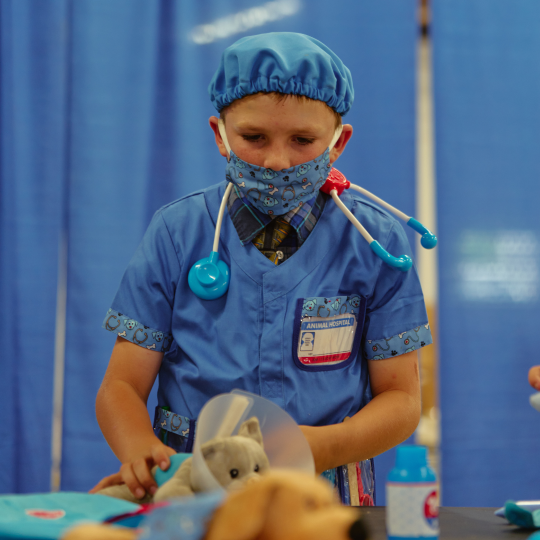 A child dressed in blue scrubs with a mask and stethoscope plays veterinarian, examining a stuffed animal at an interactive exhibit.