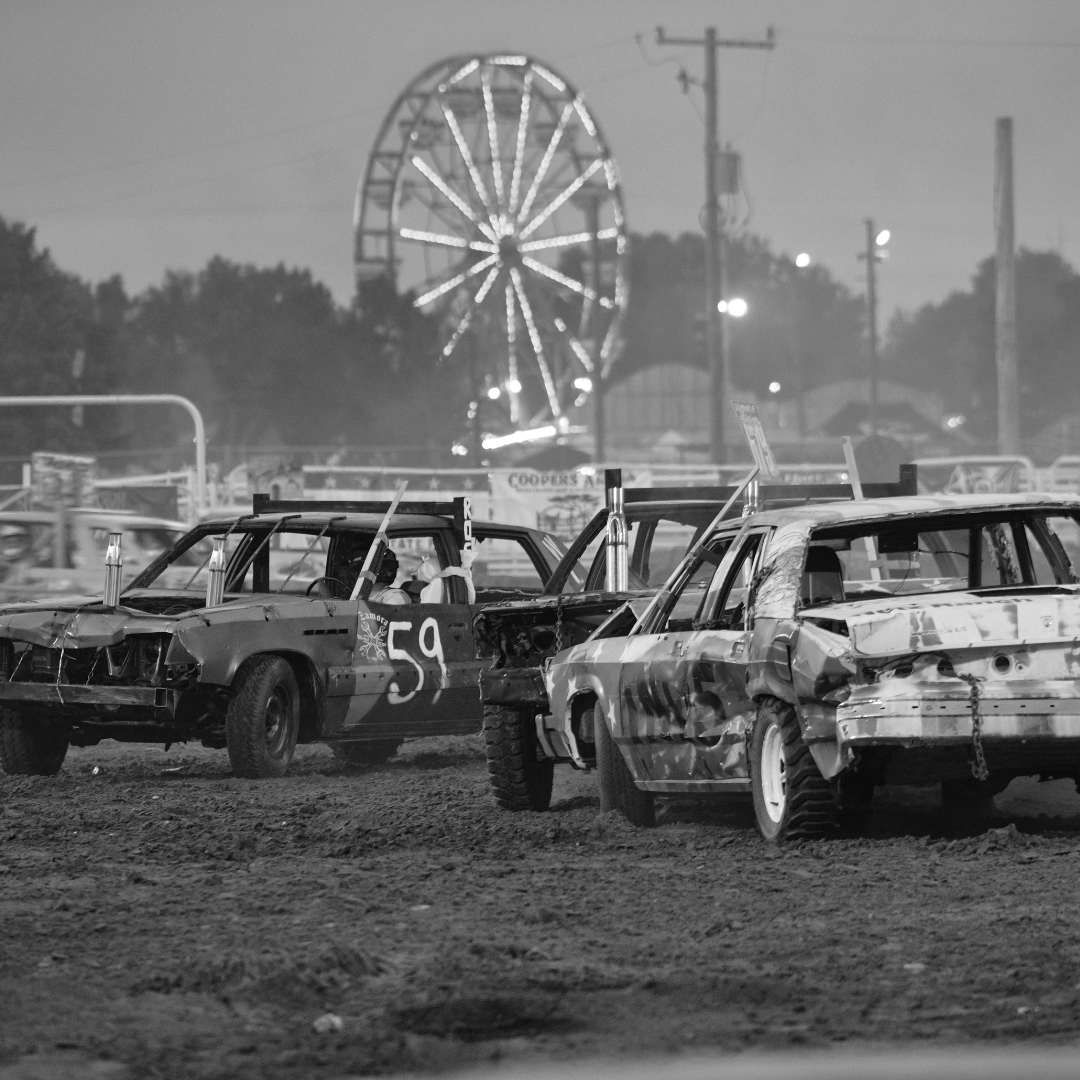 This black and white photo captures the excitement of a demolition derby at the fair, with a row of battered cars lined up in the dirt arena, their dented bodies showing signs of previous collisions. The number "59" is boldly painted on one of the vehicles, standing out against the rough, worn exterior. In the background, the glowing lights of a Ferris wheel illuminate the night sky, adding a festive touch to the gritty competition. Power lines and fairground structures frame the scene, emphasizing the blend of adrenaline-pumping action and classic fair nostalgia.