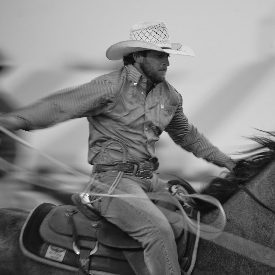 This black and white photo captures the intensity of a rodeo cowboy in action as he races across the arena on horseback. Dressed in a striped button-up shirt, jeans, and a classic cowboy hat, he has a determined expression as he swings his rope with precision. The movement of the lasso and the horse's stride are frozen in time, highlighting the speed and skill involved in the event. The details of the saddle and the cowboy's belt buckle add to the authentic Western feel of the moment. This image perfectly showcases the dedication and grit that define rodeo competition.