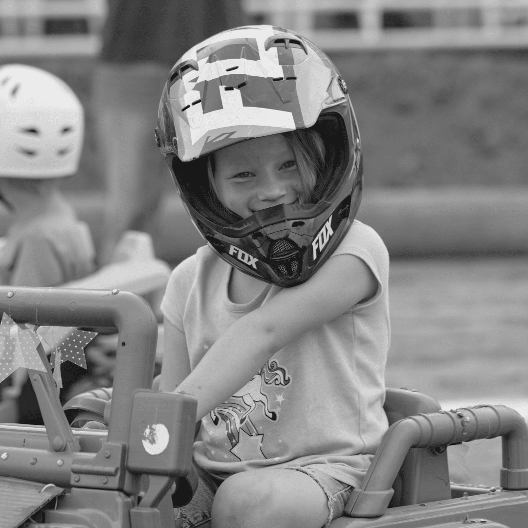 B&W Power Wheels This black and white photo captures the excitement of the Power Wheels competition at the fair, where young participants gear up to take on their own version of the demolition derby. A smiling young girl, wearing a protective helmet, sits confidently in her battery-powered vehicle, ready for action. Her vehicle is decorated with fun details, including small balloons attached to the sides, which are the targets in this playful yet competitive event. In the background, another child wearing a helmet is focused on their own ride, adding to the atmosphere of friendly competition and fun. This image perfectly showcases the joy and excitement of kids getting their first taste of derby action in a safe and thrilling way.