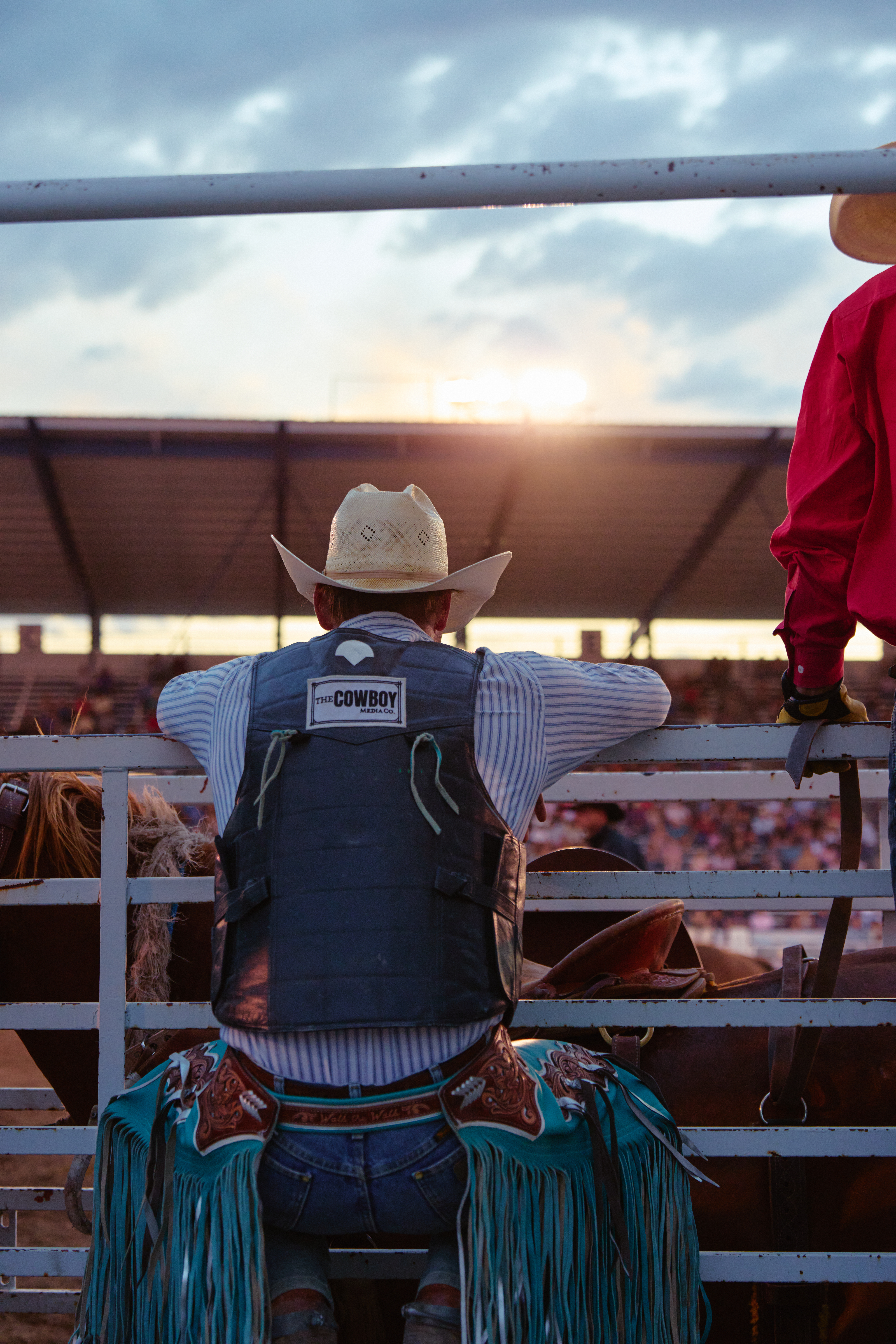 A bull rider wearing a white hat, blue chaps, and protective vest leans on a fence from horseback, watching a rodeo arena at sunset as a crowd fills the grandstands behind him.