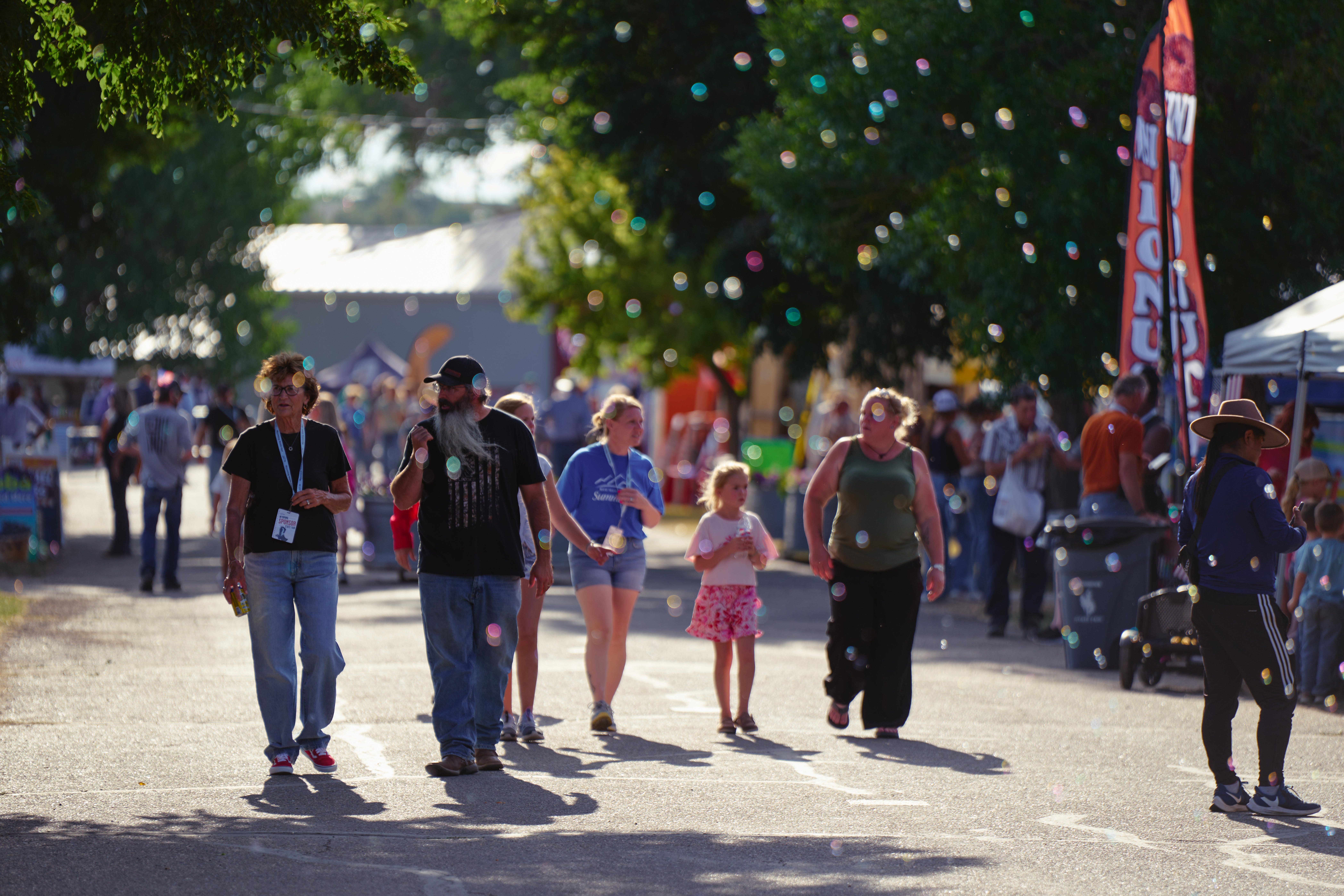 ### **Image 24 – Fairgoers on the Midway** **Alt Text:** Fairgoers of all ages walk along a tree-lined midway at the Wyoming State Fair as colorful bubbles float through the air in the evening sunlight.